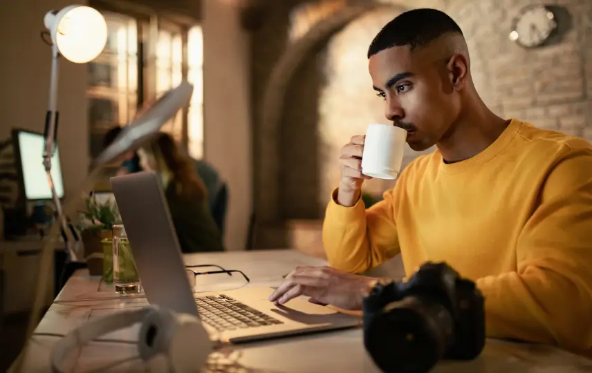Young man in yellow sweater sips coffee, focused on laptop. Camera, headphones on desk in warm lit room.