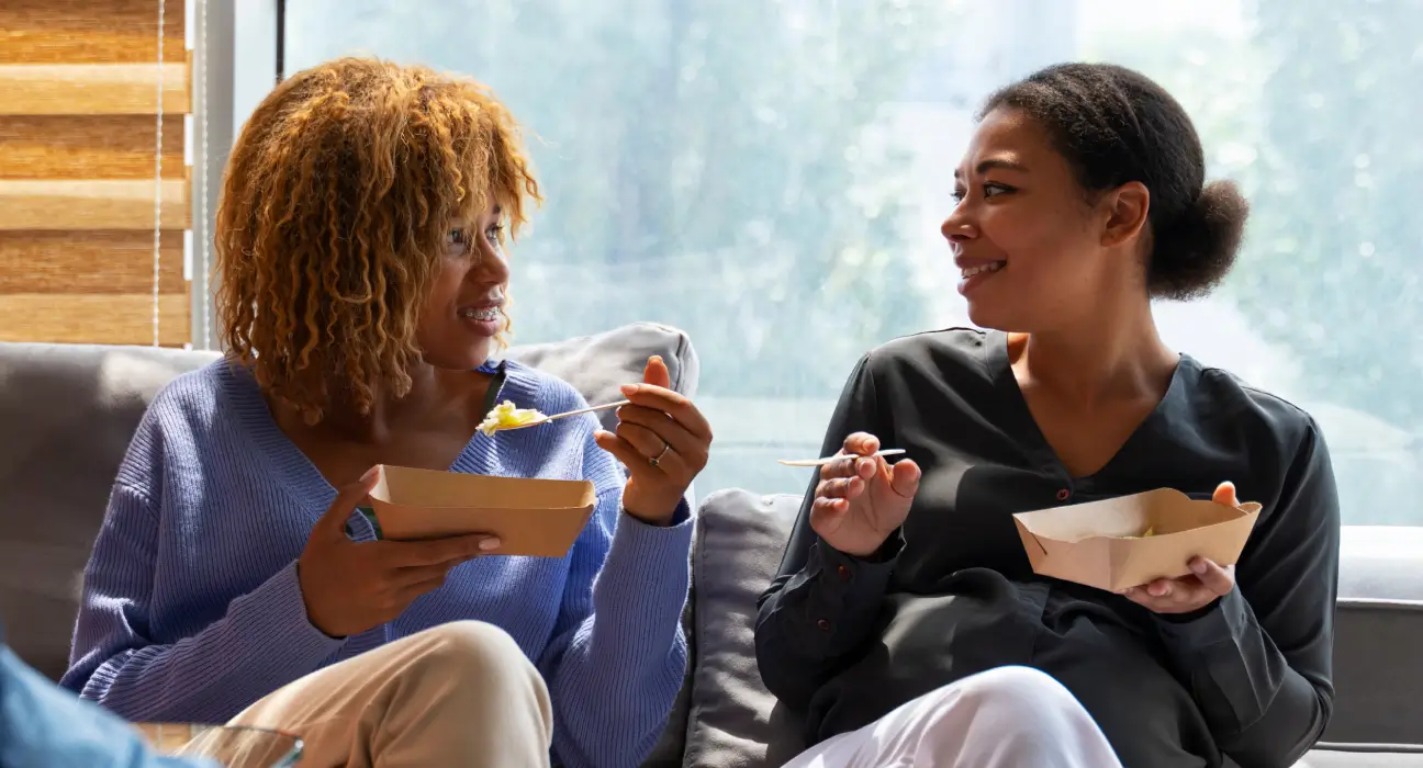 Two smiling women relax on a gray sofa, eating light meals from small paper trays and engaging in friendly conversation.