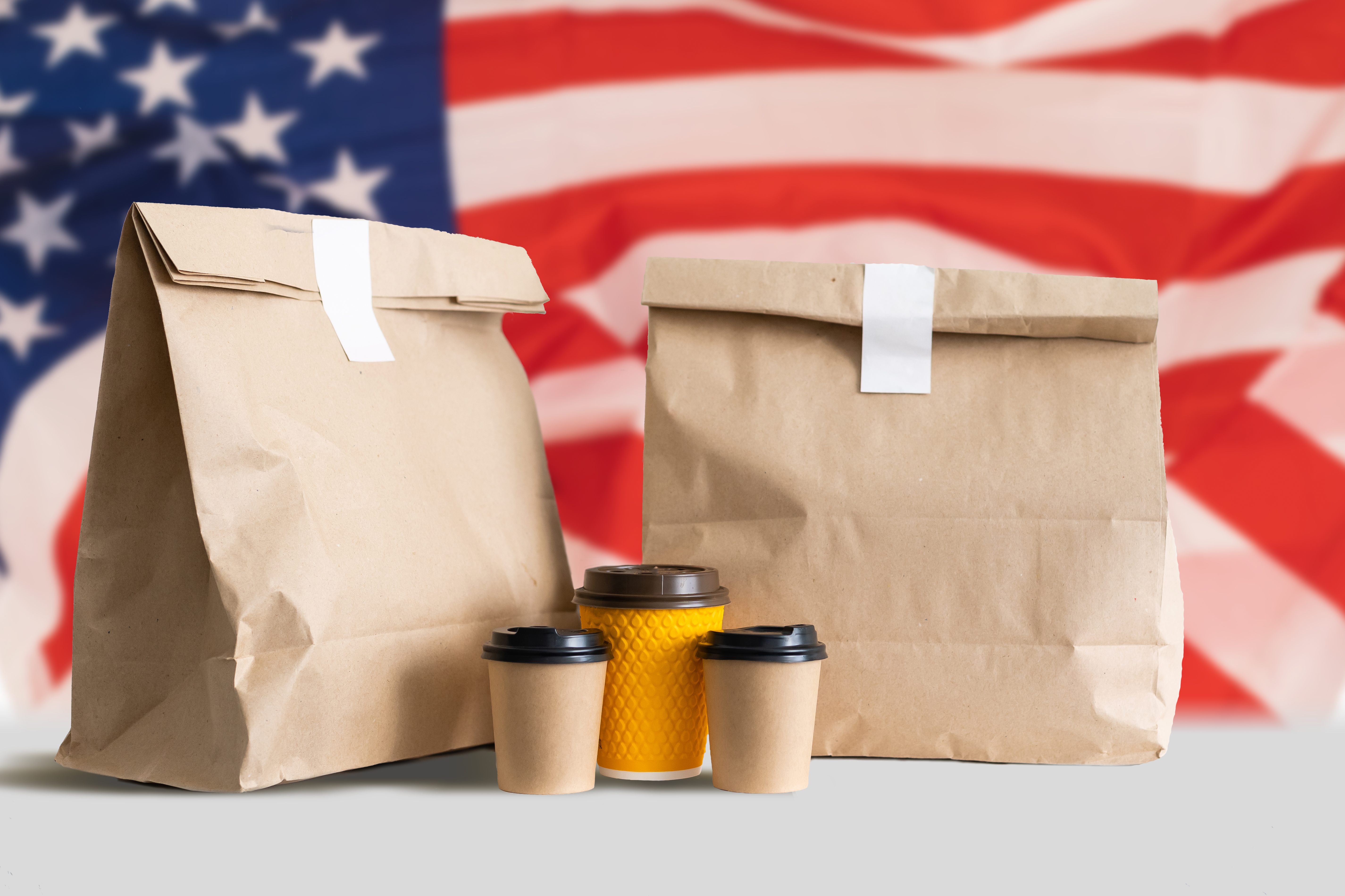 Two brown paper takeout bags and three coffee cups against a blurred American flag, symbolizing U.S. food and beverage packaging.